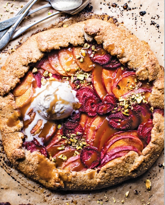 A rustic fruit galette topped with vanilla ice cream, caramel drizzle, and chopped pistachios rests on parchment paper, served comfort-food style with spoons close at hand—a scene inspired by the cozy signature touch of Tieghan Gerard as featured in the Half Baked Harvest Cookbook by Penguin Random House.
