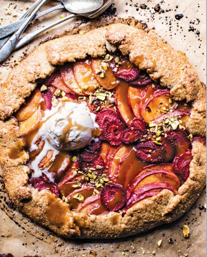 A rustic fruit galette topped with vanilla ice cream, caramel drizzle, and chopped pistachios rests on parchment paper, served comfort-food style with spoons close at hand—a scene inspired by the cozy signature touch of Tieghan Gerard as featured in the Half Baked Harvest Cookbook by Penguin Random House.