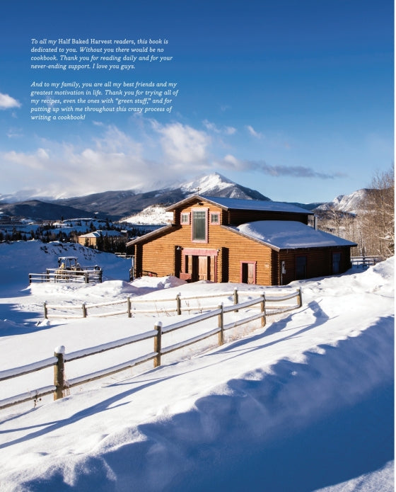 A wooden house nestled in a snowy landscape with a wooden fence out front, majestic mountains behind, and a clear blue sky overhead—an inviting scene reminiscent of the cozy settings found in the Half Baked Harvest Cookbook from Penguin Random House, where comforting recipes bring warmth to every meal.