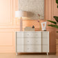 The Essery Dresser Collection by Made Goods, a modern white three-drawer dresser with soft-close glides, stands against a peach wall topped with a lamp and décor; a plant sits to the right—an elegant furniture piece.