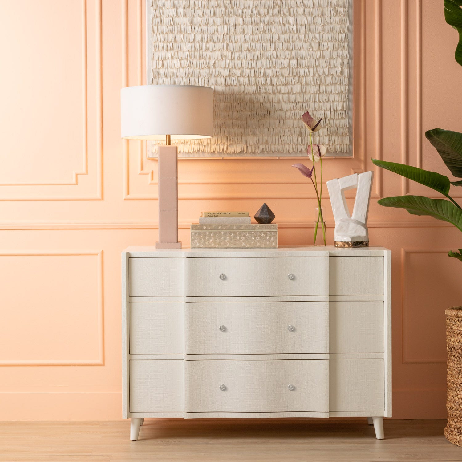 The Essery Dresser Collection by Made Goods, a modern white three-drawer dresser with soft-close glides, stands against a peach wall topped with a lamp and décor; a plant sits to the right—an elegant furniture piece.
