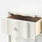 A close-up of a white textured nightstand from the Made Goods Essery Nightstand Collection features faux raffia, a partially open drawer revealing a light wood interior, and a round silver knob.