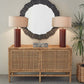 The Made Goods Isla Buffet serves as an elegant dining room essential, topped with two brown table lamps and a decorative box, while a round dark-framed mirror hangs above and a leafy plant stands nearby.