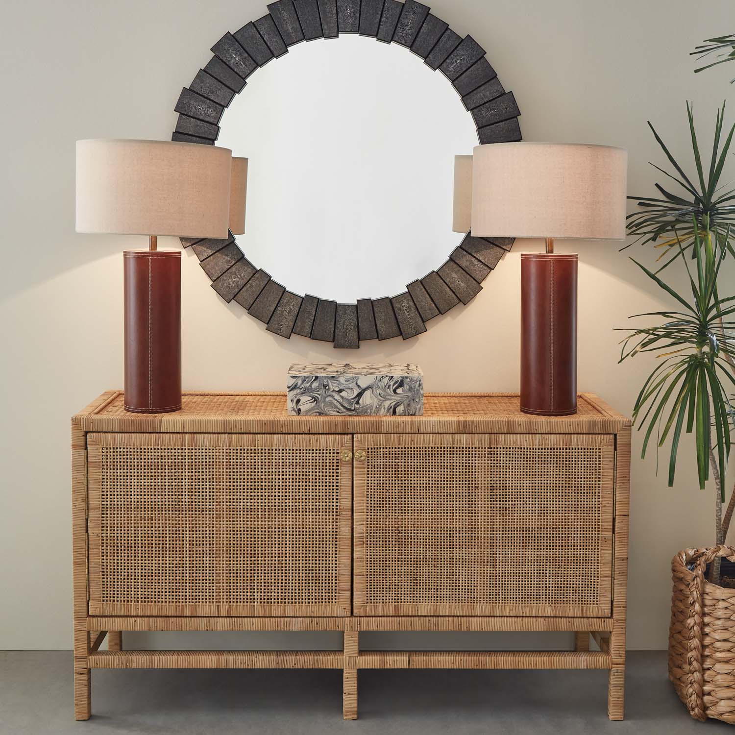 The Made Goods Isla Buffet serves as an elegant dining room essential, topped with two brown table lamps and a decorative box, while a round dark-framed mirror hangs above and a leafy plant stands nearby.