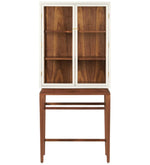 The Made Goods Jamison Cabinet features glass-paneled doors, three shelves, and stands on a matching wooden base with walnut legs and an open lower section.