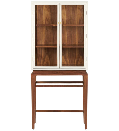 The Made Goods Jamison Cabinet features glass-paneled doors, three shelves, and stands on a matching wooden base with walnut legs and an open lower section.