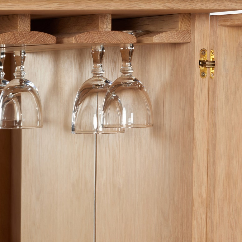 Two upside-down wine glasses hang from a wooden rack inside the Vanna Bar Cabinet by Made Goods, featuring a light oak veneer finish.