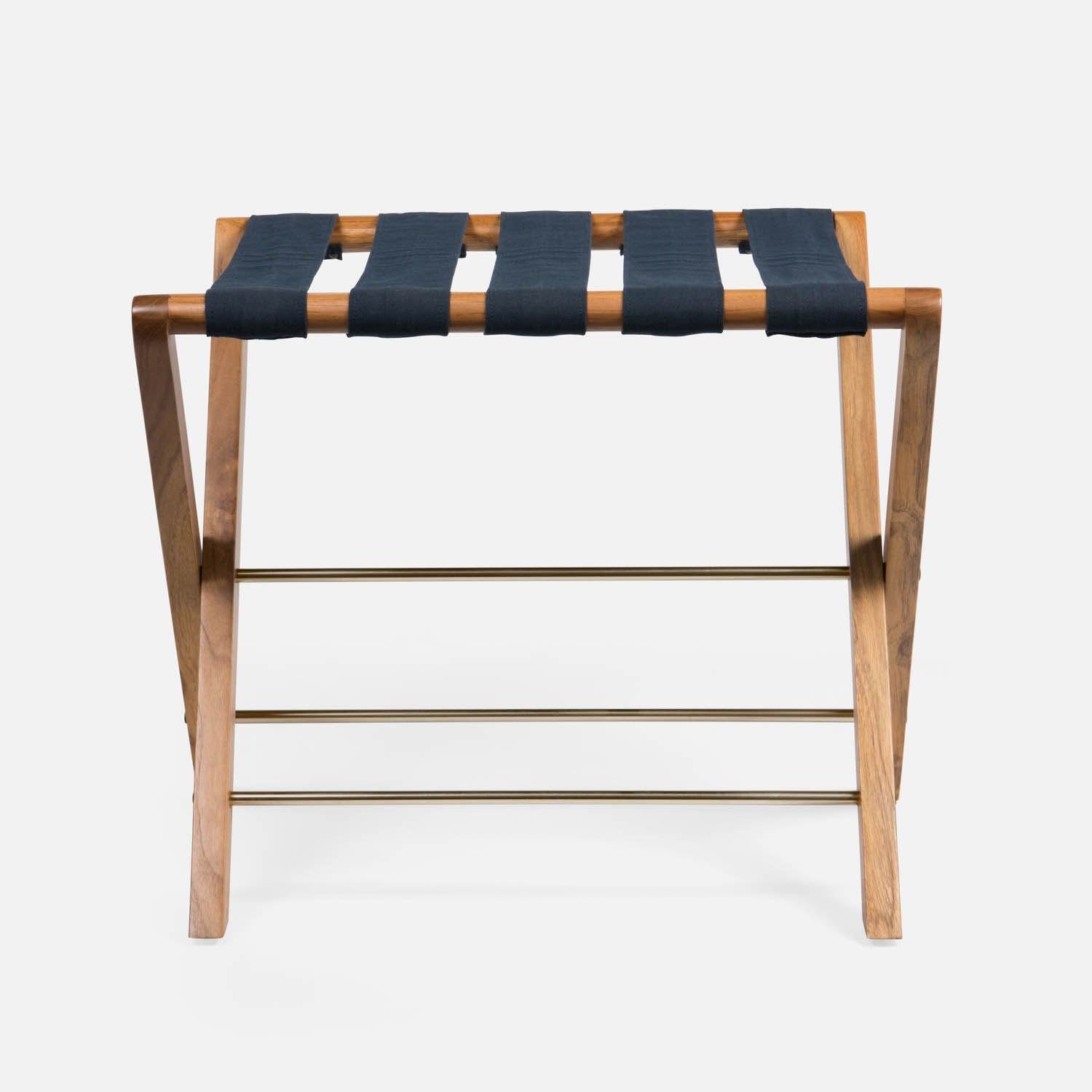 The Made Goods Walvia Teak and Navy Luggage Rack features crossed legs, brass support bars, and a top of navy canvas straps, shown against a white background.