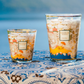 Two Baobab Collection Cities Côte d’Azur Candles, by Baobab, with elegant screen-printed designs, are displayed on a blue and white patterned tablecloth against a softly blurred outdoor background.