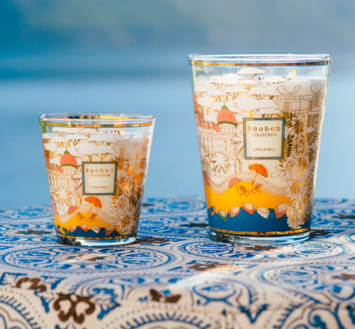 Two Baobab Collection Cities Côte d’Azur Candles, by Baobab, with elegant screen-printed designs, are displayed on a blue and white patterned tablecloth against a softly blurred outdoor background.