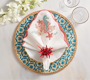 The Kim Seybert Marina Napkin Set adds ocean-inspired elegance to your table, especially when paired with woven placemats, blue and gold plates, coral napkin rings, and chic glassware.