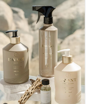 Four L'Avant Collective Ambre Santal Collection products, with cardamom, amber, and cedar notes, are arranged on a reflective surface with natural light and driftwood, evoking the elegance of luxury perfume.
