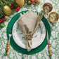 A festive holiday table is set with a deer plate, a beige napkin accented by the Kim Seybert Gem Wreath Napkin Ring Set, bamboo-handled cutlery, two glasses, and greenery with berries and gold ornaments.