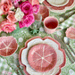 A floral-themed table is set with Bordallo Pinheiro’s Pink Cabbage Leaf Dinnerware and Serveware Collection, silver cutlery, a pink mug, glass, and a vase of pink flowers atop a green patterned tablecloth.