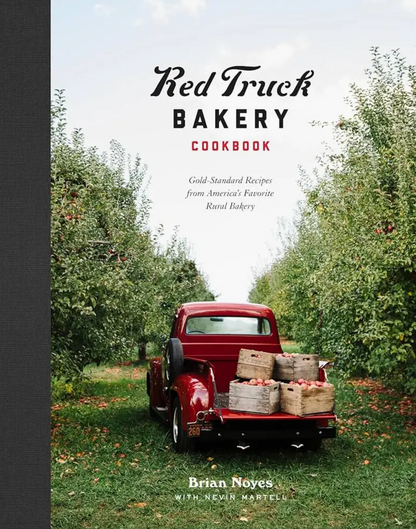 A red vintage truck filled with baskets of apples sits between rows of apple trees, while “Red Truck Bakery Cookbook” by Penguin Random House—featuring delicious southern baking recipes—appears as the title on the book’s cover.