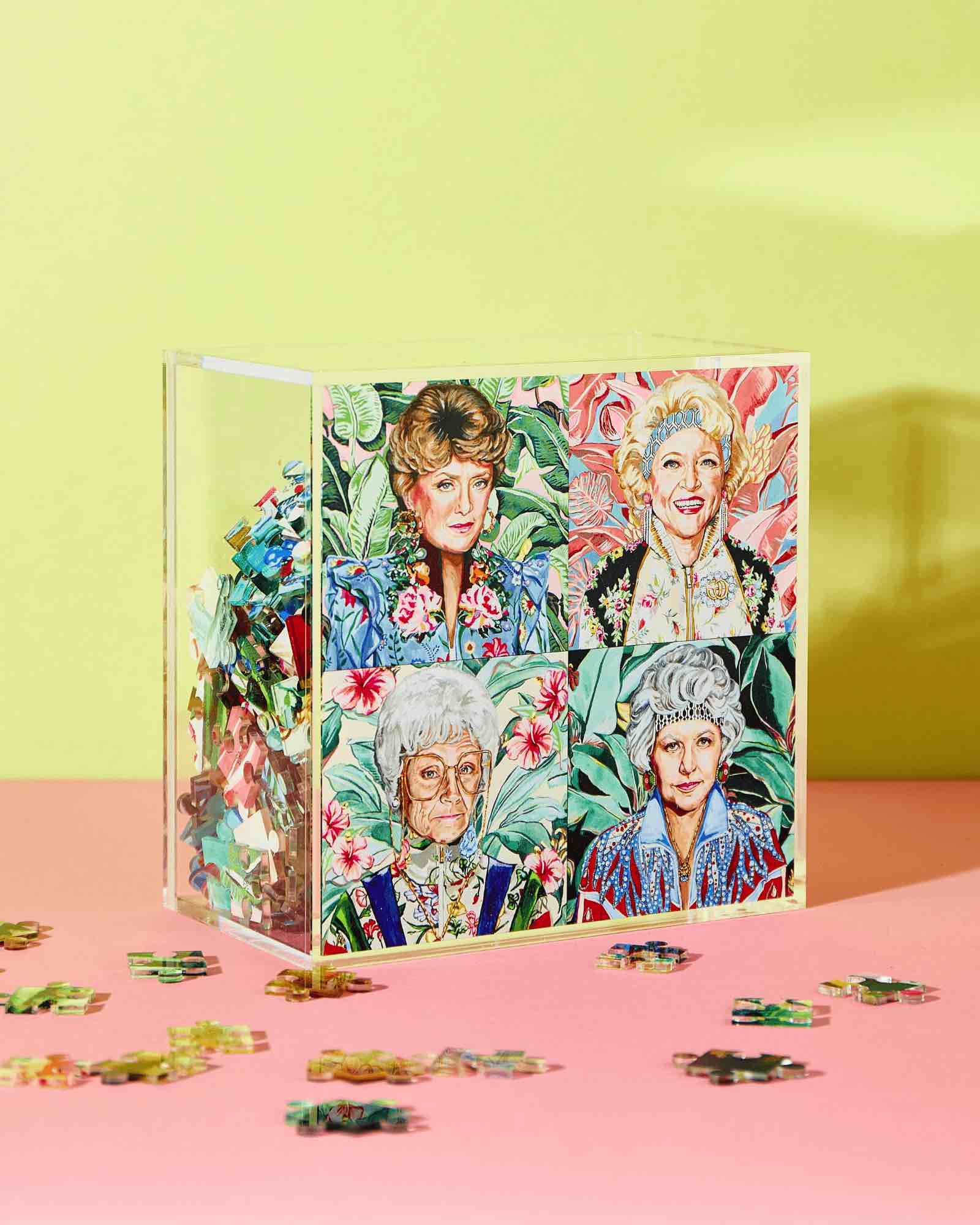 The Acrylic Puzzle by Tart by Taylor features four older women with floral backgrounds, displayed in a clear acrylic box. Loose pieces on a pink surface add charm, making it a stylish home decor piece.