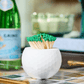 The Tart by Taylor Golf Match Striker Collection features a white textured container holding green-tipped wooden matches, making a stylish accessory for your tray alongside books, a glass bottle, and a glass.