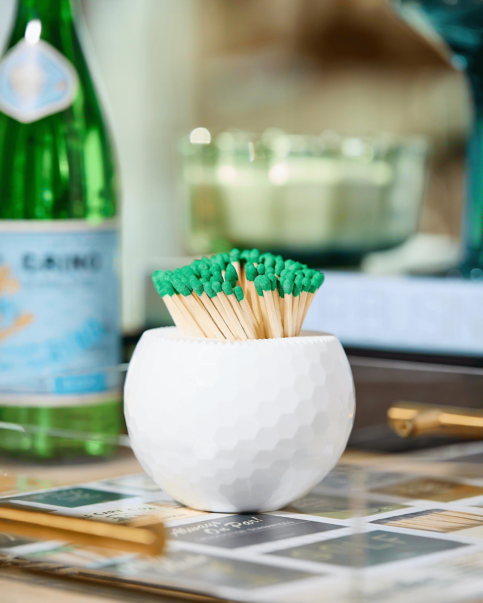 The Tart by Taylor Golf Match Striker Collection features a white textured container holding green-tipped wooden matches, making a stylish accessory for your tray alongside books, a glass bottle, and a glass.