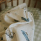 The Gooselings Transit Treasures Swaddle, a cream-colored 100% cotton baby blanket featuring blue vintage car illustrations, is draped over a green and white checkered crib mattress.