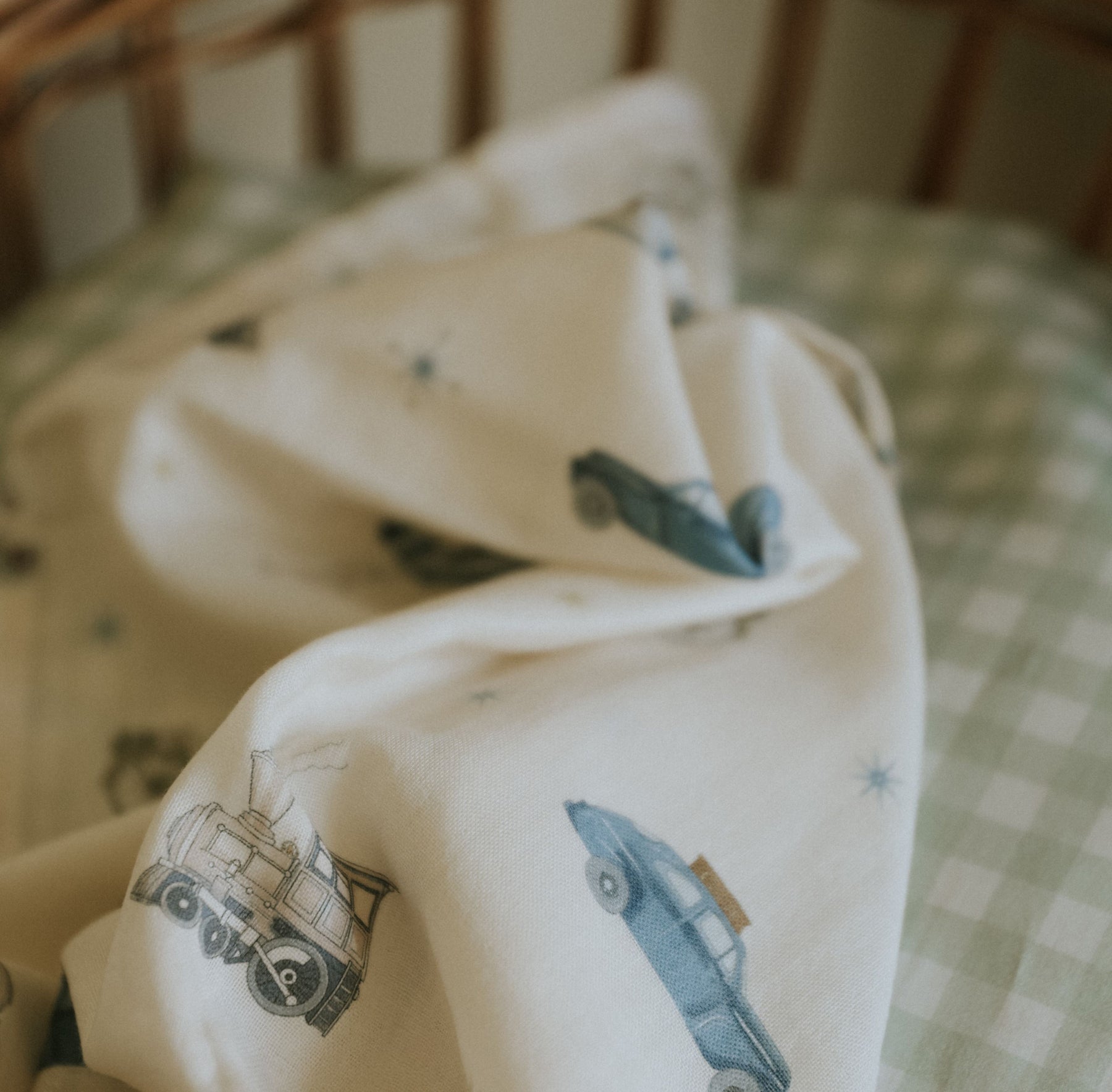The Gooselings Transit Treasures Swaddle, a cream-colored 100% cotton baby blanket featuring blue vintage car illustrations, is draped over a green and white checkered crib mattress.