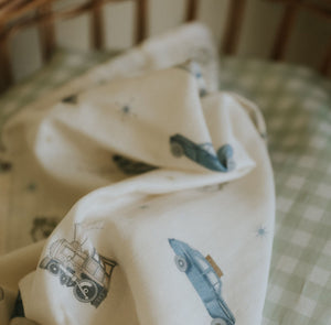 The Gooselings Transit Treasures Swaddle, a cream-colored 100% cotton baby blanket featuring blue vintage car illustrations, is draped over a green and white checkered crib mattress.