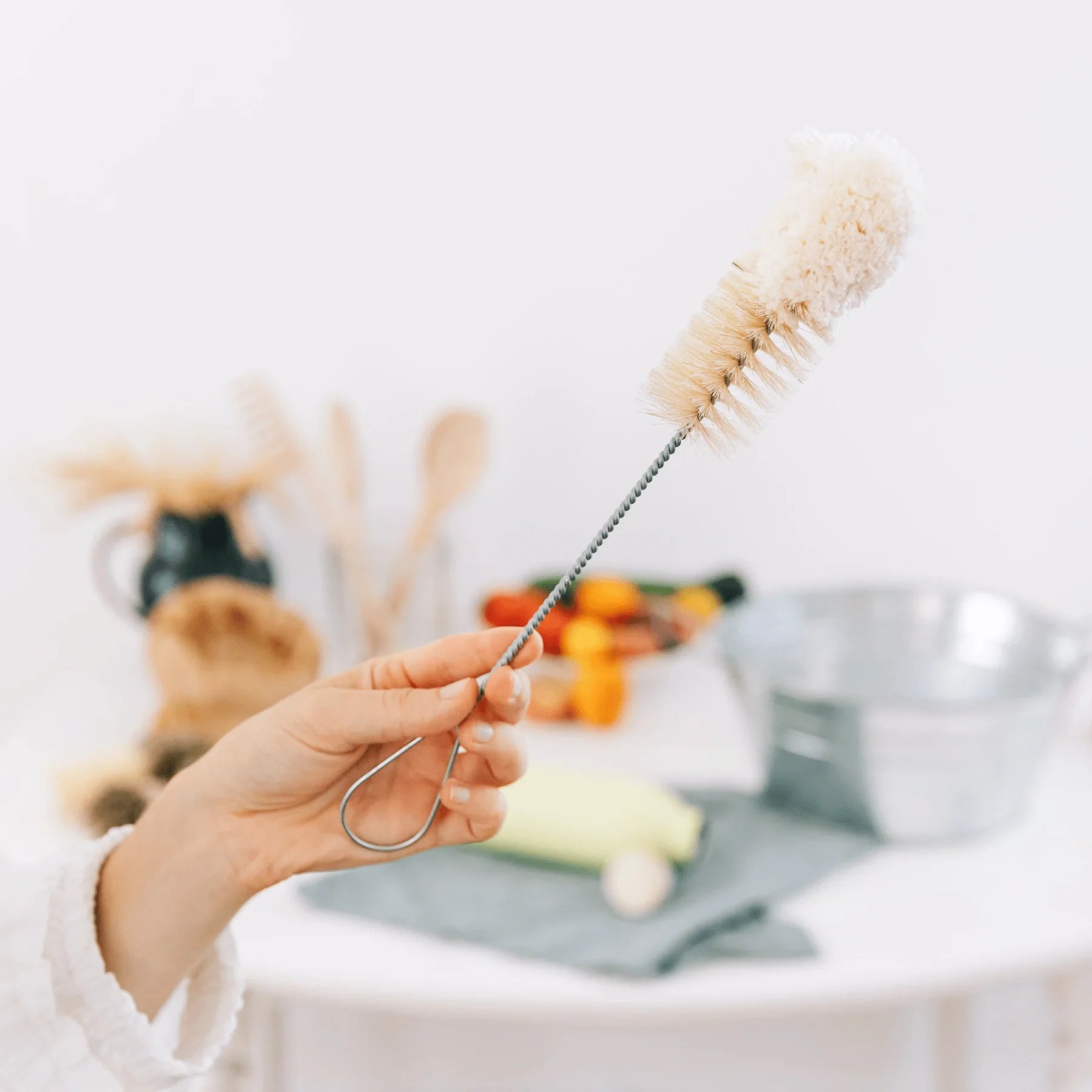 A hand holds the Be Home Bistro Flute Cleaning Brush, ideal for cleaning small glassware such as champagne flutes, with blurred kitchen utensils and cleaning supplies in the background.
