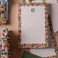 The Gracie Notepad by Rifle Paper Co. is an illustrated notepad with a scalloped floral border and a small blue and green flower at the top, featuring tear-off sheets, shown on a beige fabric surface.