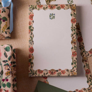 The Gracie Notepad by Rifle Paper Co. is an illustrated notepad with a scalloped floral border and a small blue and green flower at the top, featuring tear-off sheets, shown on a beige fabric surface.