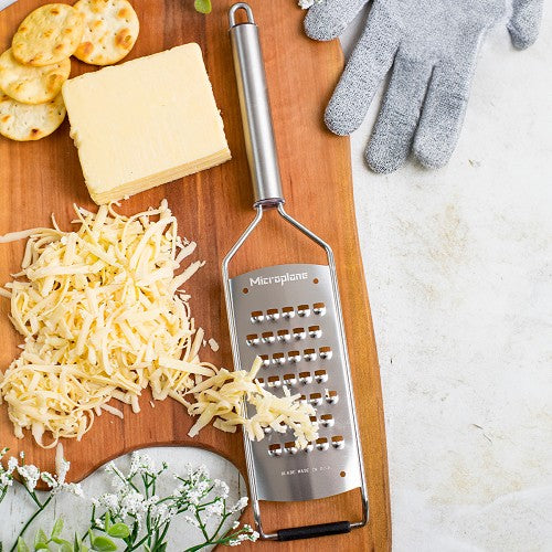 The Microplane Professional Series 2.0 Grater Collection is shown on a wooden cutting board with grated cheese, a cheese block, crackers, and gray gloves.