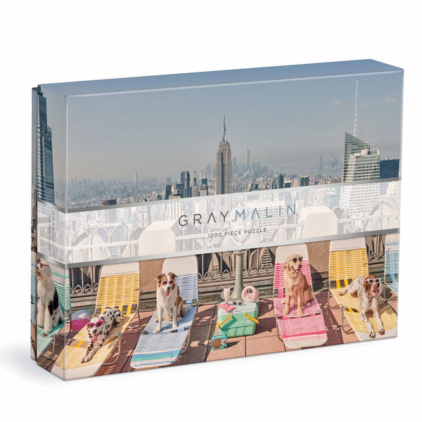 Experience the charm of Chronicle Books' Gray Malin 1000 Piece Puzzle, "The Dogs of New York City," featuring a rooftop scene with five dogs relaxing on sun loungers against a stunning cityscape and towering skyscrapers.