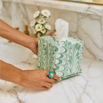 Someone sets a green and white tissue box from the Marble Bath Collection by Weezie Towels on a marble countertop near a sink and a vase of white flowers.