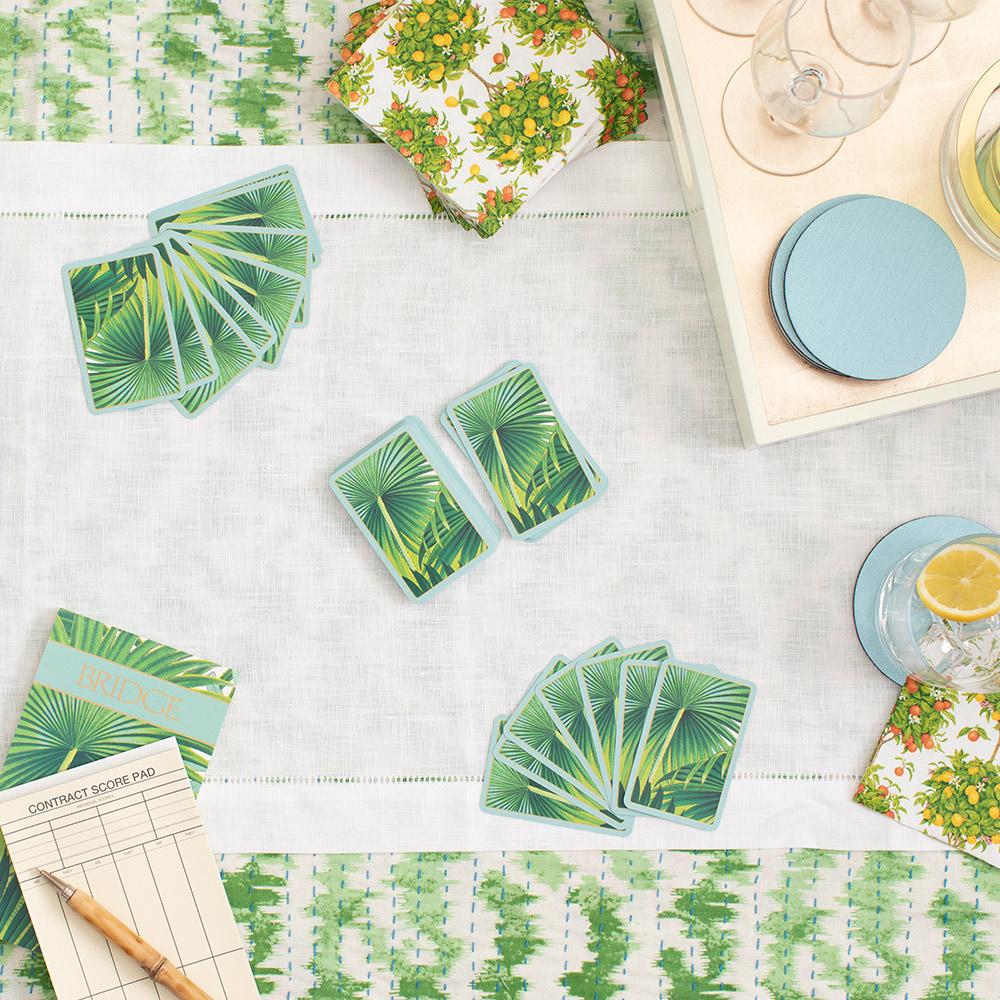 Overhead view of a table set for a card game featuring the Caspari Palm Fronds Bridge Gift Set with tropical-themed playing cards, bridge score pads, a pencil, drinks with lemon, blue coasters, and a floral napkin.