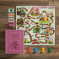 The WS Game Company Candy Land Vintage Bookshelf Edition, ideal for a child's first game, features a vibrant game board, four character pieces, cards, and instructions all neatly arranged in a charming pink book-shaped box and elegantly displayed on a wooden surface.