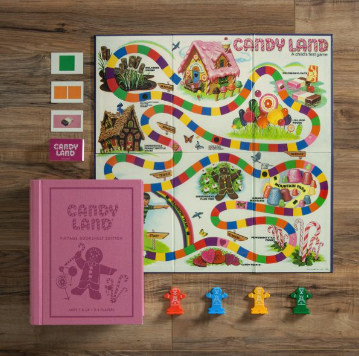 The WS Game Company Candy Land Vintage Bookshelf Edition, ideal for a child's first game, features a vibrant game board, four character pieces, cards, and instructions all neatly arranged in a charming pink book-shaped box and elegantly displayed on a wooden surface.