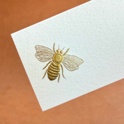 The Printery Bee Place Card Set features bone white cards with an embossed gold bee illustration on a brown background—elegant tented place cards perfect for your special event.