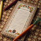 The Harvest Gingham Market Pad by Rifle Paper Co., featuring charming floral and vegetable designs, sits atop a patterned tablecloth surrounded by colorful vegetable-shaped pens—ideal for organizing your Thanksgiving grocery shopping or attaching to your fridge with its magnet for easy access.