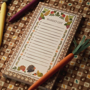 The Harvest Gingham Market Pad by Rifle Paper Co., featuring charming floral and vegetable designs, sits atop a patterned tablecloth surrounded by colorful vegetable-shaped pens—ideal for organizing your Thanksgiving grocery shopping or attaching to your fridge with its magnet for easy access.