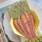 A place setting from the Farmer's Market Collection showcases a yellow bowl with a napkin and the Hester & Cook Carrots Table Accent labeled "Peggy," set on a plate with utensils beside it.