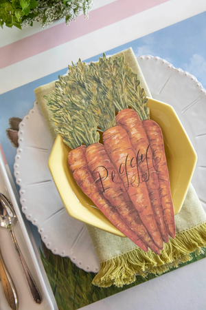 A place setting from the Farmer's Market Collection showcases a yellow bowl with a napkin and the Hester & Cook Carrots Table Accent labeled "Peggy," set on a plate with utensils beside it.