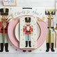 Holiday table decor from the Hester & Cook Nutcracker Collection featuring a nutcracker-themed paper placemat, gold-rimmed pink plate, and a nutcracker-patterned paper napkin with a “Cheers to You!” banner.