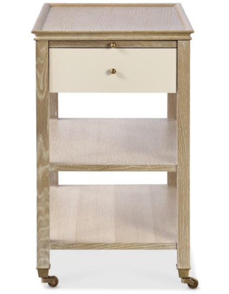 The Highland House Alexander Narrow Bedside Table features two open shelves, a drawer, and four caster wheels for easy mobility, offering stylish bedside storage in a blonde cerused finish.
