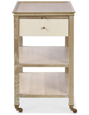 The Highland House Alexander Narrow Bedside Table features two open shelves, a drawer, and four caster wheels for easy mobility, offering stylish bedside storage in a blonde cerused finish.