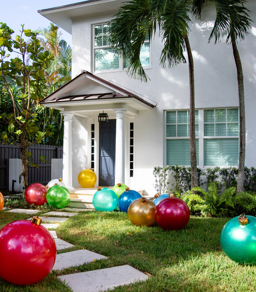 A white two-story house showcases Holiball Giant Inflatable Ornaments from the Holiball brand scattered across the lawn in the front yard, creating a festive atmosphere. Nearby palm trees sway gently, merging tropical vibes with the holiday decorations.