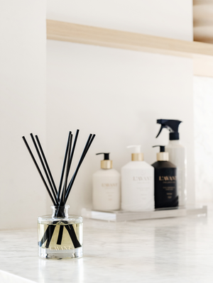 A Fresh Linen Reed Diffuser by L'Avant Collective sits on a marble countertop, accompanied by four bottles of cleaning and personal care products in the background.