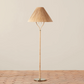 The Delphine Floor Lamp by Selamat features a tall, slender wooden stand with a handcrafted rattan shade and subtle brass accents on its round metal base, placed on a tiled floor against a plain beige background.