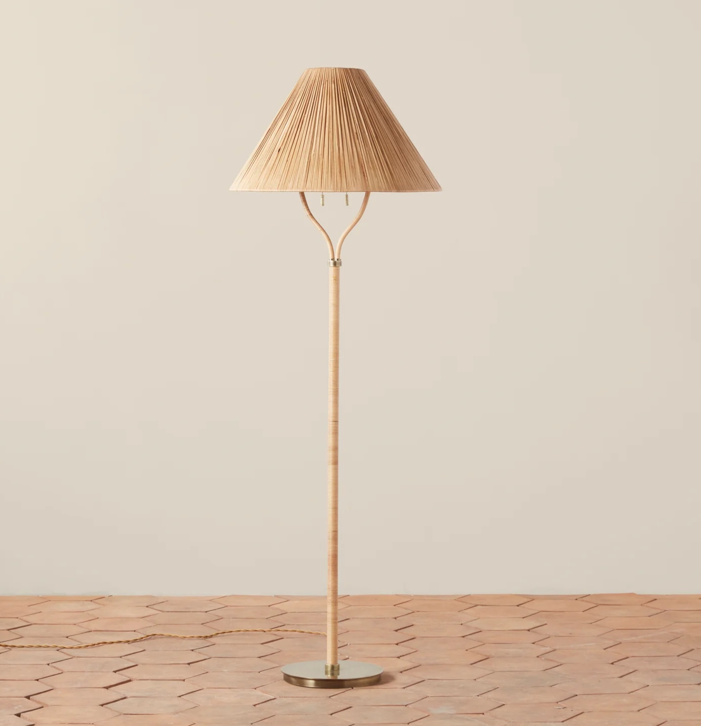 The Delphine Floor Lamp by Selamat features a tall, slender wooden stand with a handcrafted rattan shade and subtle brass accents on its round metal base, placed on a tiled floor against a plain beige background.
