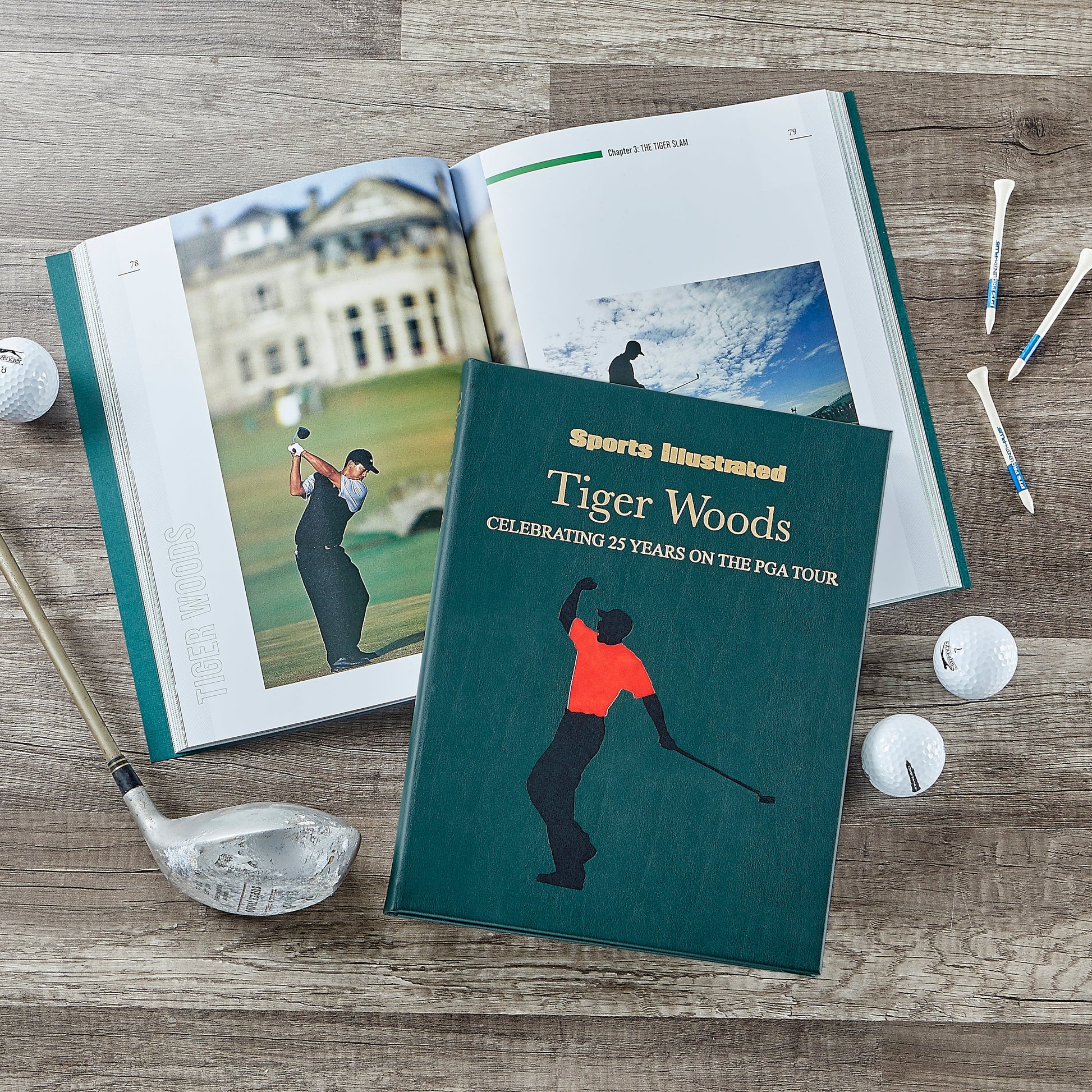 The Graphic Image book "Tiger Woods: Celebrating 25 Years on the PGA Tour" rests on a wooden surface with a golf club, balls, tees, and an open page showing legendary golfer Tiger Woods.