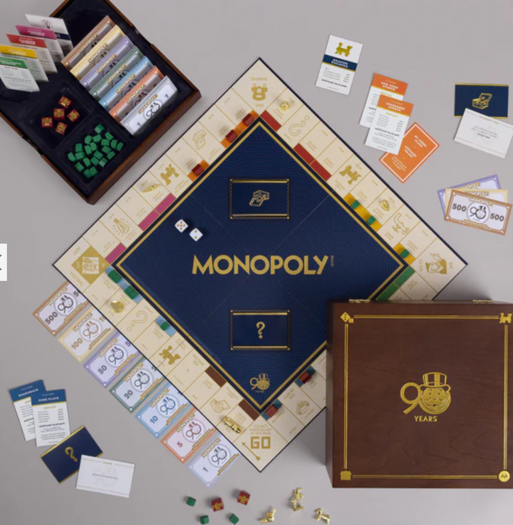 The WS Game Company Monopoly 90th Anniversary Edition features a dark blue and gold board, cards, play money, tokens, dice, and a wooden “90 Years” box—all displayed on a light surface.