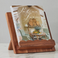 The etúHOME Scalloped Wood Book Stand, made from sustainable acacia wood, displays an open "Patina Farm" cookbook on a marble surface set against a light gray wall.