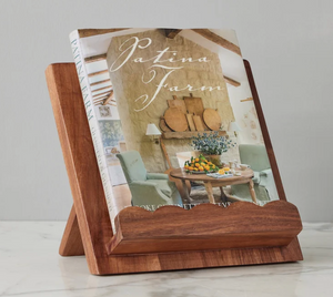 The etúHOME Scalloped Wood Book Stand, made from sustainable acacia wood, displays an open "Patina Farm" cookbook on a marble surface set against a light gray wall.